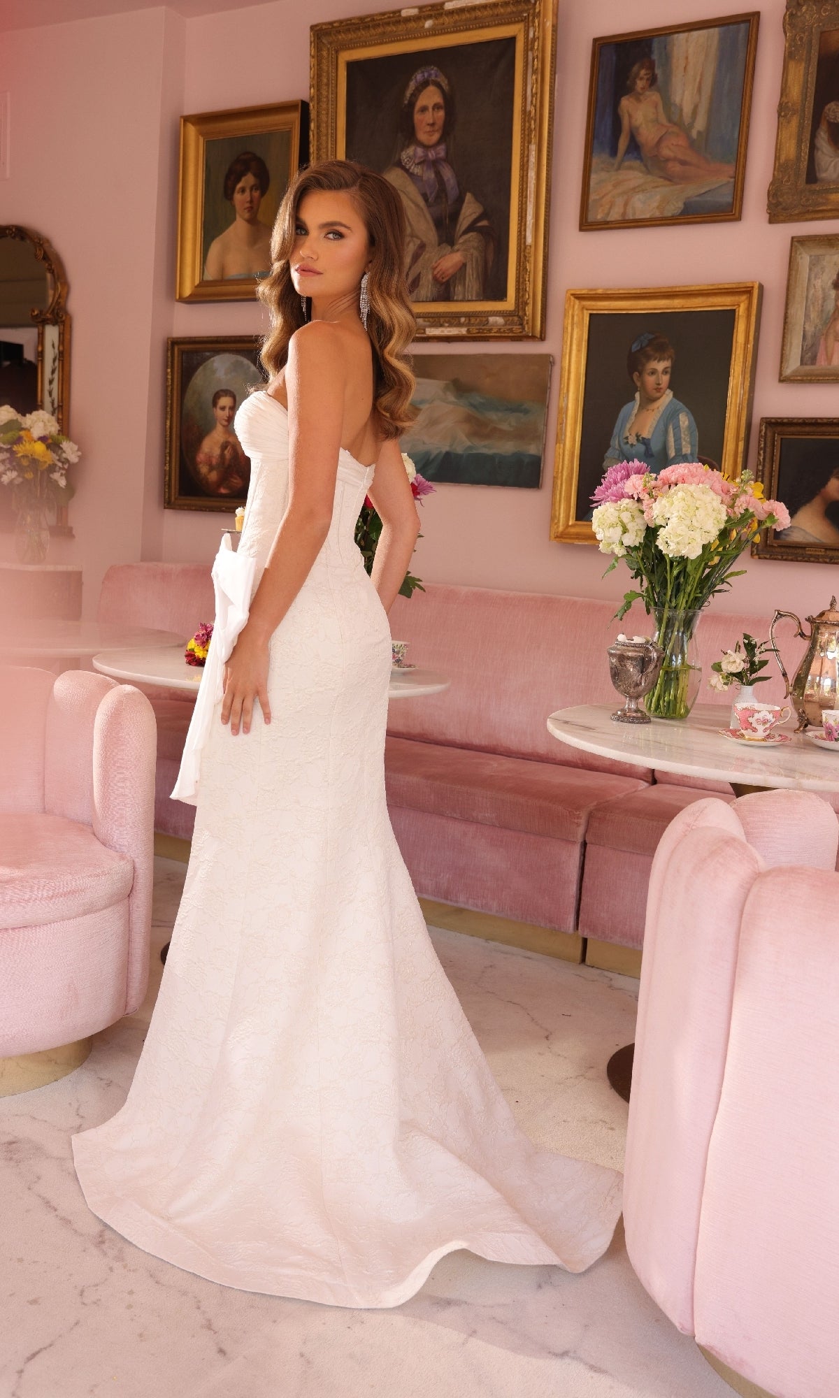 A woman wearing the Ava Presley 40099 long lace prom dress with a bow stands in a pink room with antique portraits, pink velvet chairs, and a table topped with a flower bouquet.
